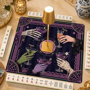 May include: A square mahjong mat featuring a dark purple and black striped background with spiderweb corners and illustrations of various hands holding cocktail glasses. A gold lamp sits in the center, surrounded by mahjong tiles with Chinese characters.