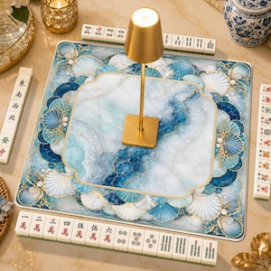 Puede incluir: Un tapete cuadrado de mahjong con un patrón de mármol azul y blanco con motivos de conchas y corales. Una lámpara dorada se sitúa en el centro. Cuatro filas de fichas de mahjong con caracteres y símbolos rodean el perímetro del tapete.