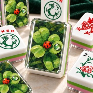 May include: A collection of rectangular Mahjong tiles featuring white surfaces with green and pink layered bases. Some tiles display painted green clover leaves with a red ladybug, while others show a red dragon, a green dragon with the text JOKER, or a flower.