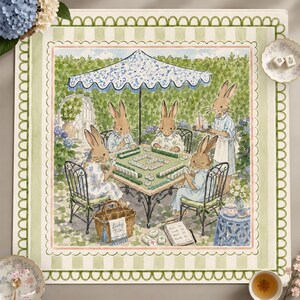 May include: A square fabric mat featuring a watercolor illustration of four rabbits playing mahjong under a blue and white umbrella in a garden. The border has a green scalloped pattern on a light green striped background with floral accents.