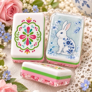May include: Three rectangular mahjong tiles with a layered design featuring a white top, a green middle band, and a translucent pink base. One tile displays a colorful floral pattern, while another features a white rabbit with blue floral accents.