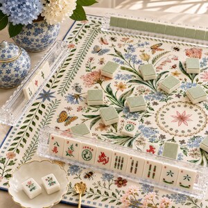 May include: A Mahjong set featuring white tiles with red, green, and black symbols, including Joker tiles, arranged in clear acrylic racks on a cream-colored mat decorated with a colorful floral, butterfly, and bee pattern.