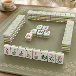 May include: A Mahjong set featuring white tiles with green and red ink illustrations of dragons, flowers, and the word Joker. The tiles have a layered design with a light green and pink base, arranged in rows and stacks on a green mat with floral borders.