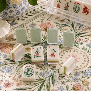 May include: A set of Mahjong tiles featuring white faces with green and red ink illustrations of flowers, dragons, and Chinese characters, alongside tiles with textured sage green backs and clear acrylic edges, arranged on a floral patterned surface.