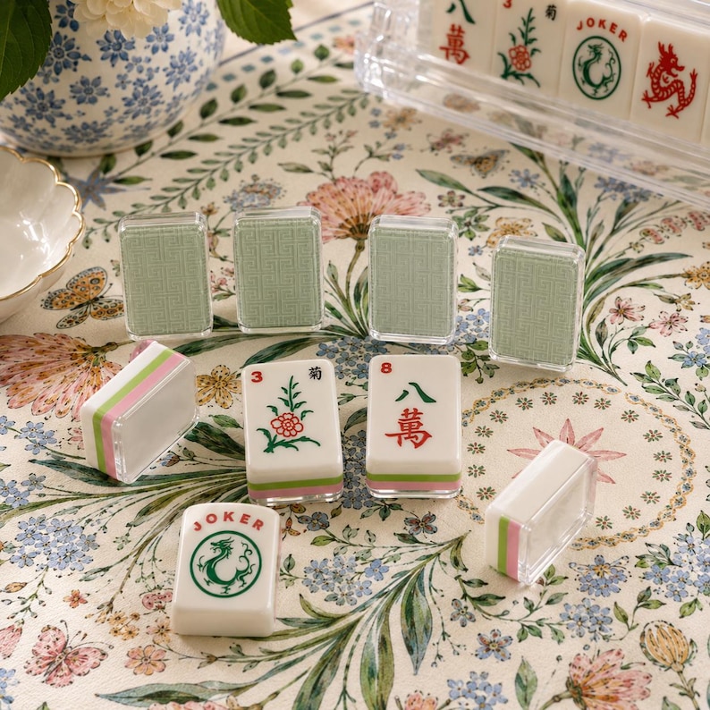 May include: A set of Mahjong tiles featuring white faces with green and red ink illustrations of flowers, dragons, and Chinese characters, alongside tiles with textured sage green backs and clear acrylic edges, arranged on a floral patterned surface.