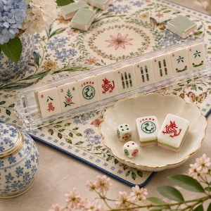 May include: A set of off-white Mahjong tiles featuring red and green ink illustrations of dragons, flowers, and numbers, displayed in a clear acrylic rack, with two loose tiles and two small white dice resting in a scalloped ceramic dish.