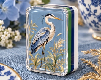 May include: A rectangular acrylic paperweight featuring a detailed illustration of a blue heron standing among golden reeds. The object has a clear front panel, a light blue background, and side edges striped in green, dark blue, and white.
