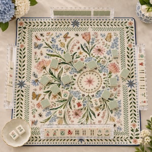 May include: A square mahjong board featuring a cream background with a colorful floral and butterfly pattern, surrounded by a blue border. The set includes clear acrylic tile racks, rectangular tiles with green backs, and tiles with red and green symbols.