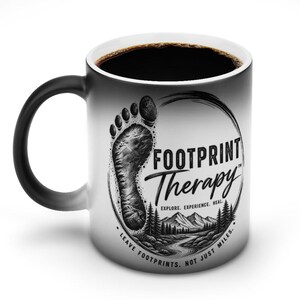Könnte beinhalten: Ein weißer Keramikbecher mit schwarz verlaufendem Henkel und Rand, verziert mit einer schwarzen Grafik eines Fußabdrucks, einer Berglandschaft und dem Text Footprint Therapy, Explore, Experience, Heal, Leave Footprints, Not Just Miles.