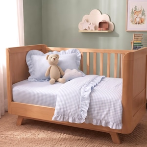 Puede incluir: Una cuna de madera clara con ropa de cama de cuadros vichy en azul claro y blanco con bordes de volantes. El juego incluye una almohada y una manta. Un oso de peluche de ganchillo beige y una pequeña almohada blanca en forma de nube.