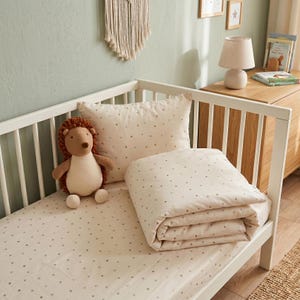 May include: A white crib bedding set featuring a pattern of small grey stars, including a fitted sheet, a pillowcase, and a folded duvet. A plush brown and cream hedgehog stuffed animal sits on the bed next to the pillow.