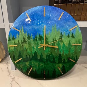 Puede incluir: Un reloj de pared redondo con una escena de bosque pintada con capas de pinos verdes que transicionan a un cielo nocturno azul estrellado. La esfera tiene marcadores de hora finos dorados y manecillas a juego.