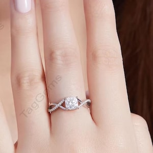 Op de afbeelding: Een zilverkleurige ring met een centrale, kussengeslepen doorzichtige steen op een band met een kruislings ontwerp. De band is versierd met kleine pavé-gezette steentjes langs de gespleten schacht, wat zorgt voor een gedetailleerde uitstraling.