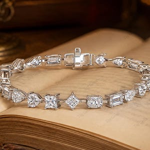 Könnte beinhalten: Ein silberfarbenes Metallarmband mit einer Reihe abwechselnder, klarer, facettierter Steine in verschiedenen Formen, darunter Rund-, Quadrat-, Smaragd- und Tropfenschliff, verbunden durch dünne Metallglieder und mit einem rechteckigen Kastenschloss.