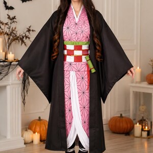 May include: A costume featuring a long black outer robe over a pink kimono with a black geometric hemp leaf pattern. It includes a white wrap skirt, a red and white checkered obi belt with a green accent, black leg wraps, and pink and black sandals.