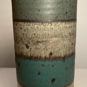May include: A cylindrical ceramic vase with a textured surface. The vase features horizontal bands of teal, beige, and dark green, with a dark brown rim. The handmade pottery has a rustic, speckled appearance.