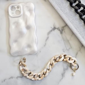 May include: An assortment of phone accessories, featuring a white textured phone case and chain straps in black, white, gold, and marbled patterns. The chains have gold-tone clasps and some have small pearl charms. A woven straw bag is also visible.