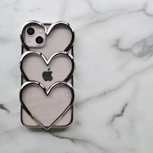 Op de afbeelding: Een transparante telefoonhoes met een zilveren hartontwerp. De hoes heeft drie harten, een binnenin de andere, en de cameralens van de telefoon is zichtbaar door het bovenste hart.