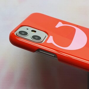 May include: A bright red phone case with a pink letter "C" printed on the back. The case has a camera lens cutout and a volume button on the side.