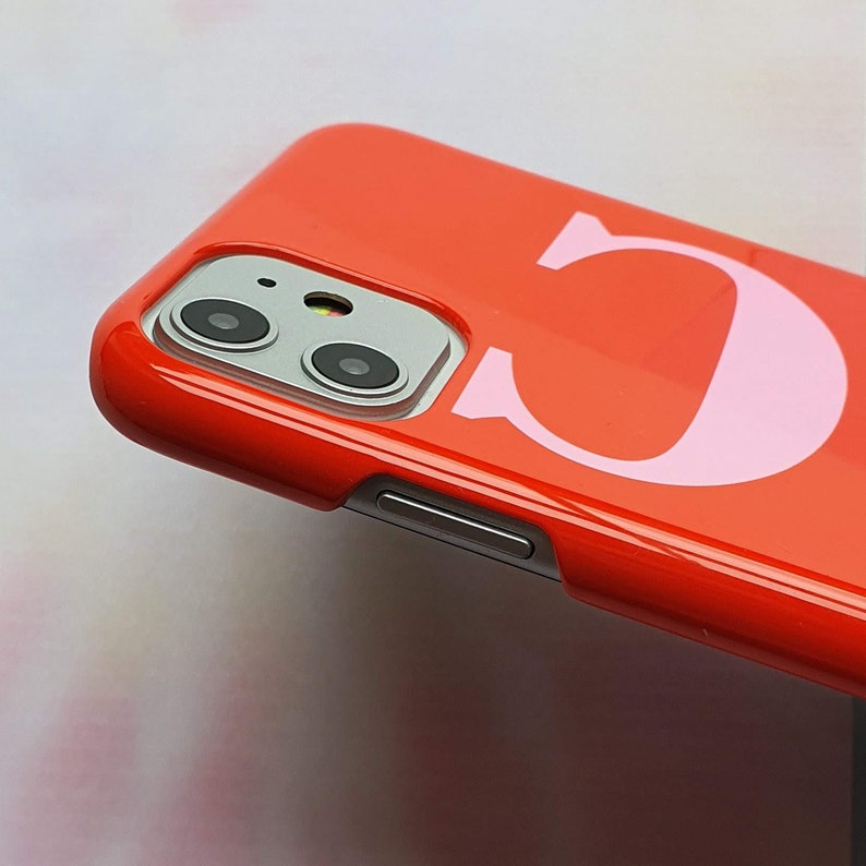 May include: A bright red phone case with a pink letter "C" printed on the back. The case has a camera lens cutout and a volume button on the side.