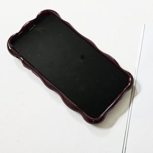 May include: A smartphone with a dark screen is encased in a burgundy-colored phone case. The case has a wavy, scalloped edge design. The phone and case are on a white surface.
