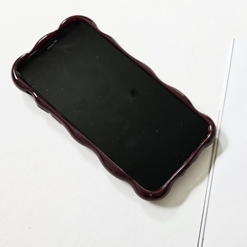 May include: A smartphone with a dark screen is encased in a burgundy-colored phone case. The case has a wavy, scalloped edge design. The phone and case are on a white surface.