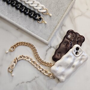 May include: An assortment of phone accessories, including two textured phone cases in white and brown, and several chain straps in black, white, gold, and a marbled pattern. Each chain has a gold clasp and a small pearl accent.