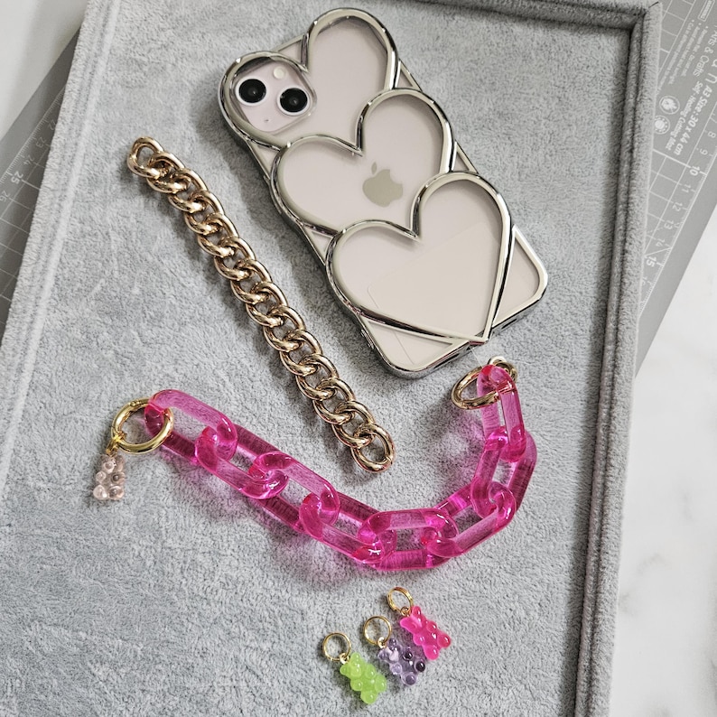 Op de afbeelding: Een transparante telefoonhoes met een drievoudig hartontwerp, een gouden ketting, een roze plastic ketting met een gouden bedel en vier gummy bear bedels in roze, paars en groen.