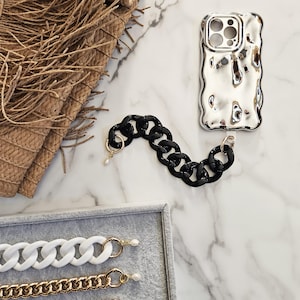 May include: Three different chain phone straps, one black, one white, and one gold. The black chain is attached to a silver phone case with a textured, wavy design.
