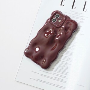 May include: A glossy, burgundy-colored phone case with a unique, wavy design. The case has a smooth, reflective surface and is designed to fit a smartphone with a dual-lens camera. The case is shown on top of a magazine.