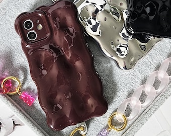 Op de afbeelding: Drie telefoonhoesjes in verschillende kleuren en stijlen. De eerste hoes is donkerbruin, de tweede is zilver en de derde is zwart. Alle drie hoesjes hebben een uniek, abstract en golvend ontwerp.