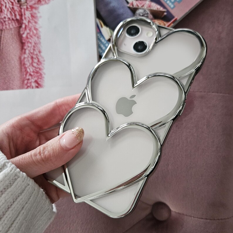 Op de afbeelding: Een transparante telefoonhoes met een zilveren metallic hartontwerp. De hoes heeft drie harten, een binnenin de andere, en een uitsparing voor de camera.