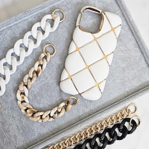 May include: A white phone case with a gold grid design, alongside several chain straps in white, marbled, gold, and black. The chains have gold-colored clasps. The items are arranged on a gray surface.