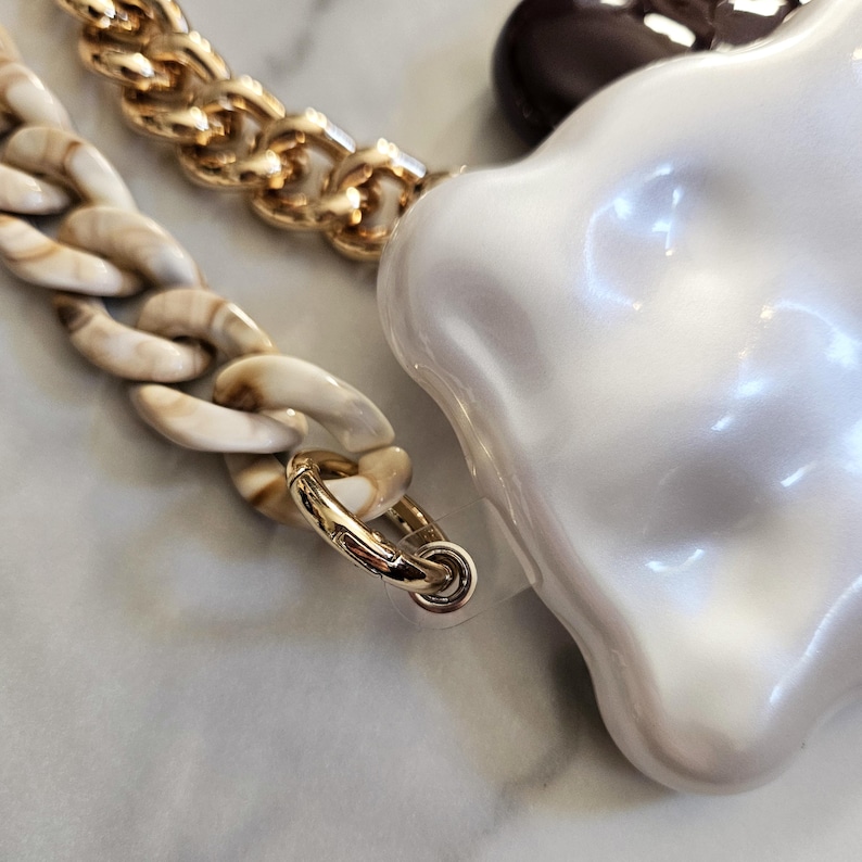 May include: Close-up of a gold chain, a marbled chain, and a large, irregular-shaped white pendant. The gold chain is linked to a marbled chain with a gold clasp. The white pendant has a pearlescent finish.