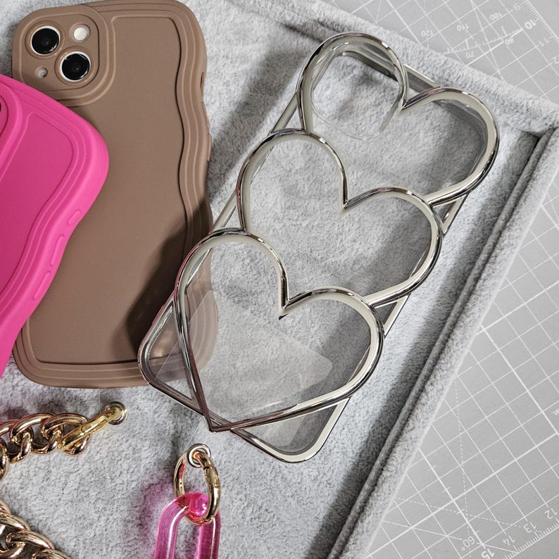 Op de afbeelding: Een transparante telefoonhoes met een zilveren hartontwerp. De hoes is gemaakt van transparant plastic en heeft een zilveren metalen hartontwerp op de achterkant. De hoes is ontworpen om te passen op een smartphone.