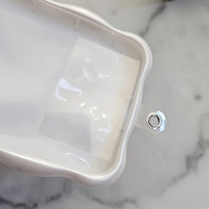 May include: A white, pearlescent phone case with a wavy, scalloped edge. The case has a clear, rectangular insert and a silver-colored metal grommet. The case is on a marble surface.