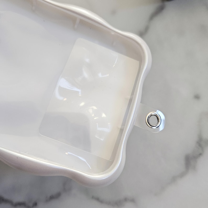 May include: A white, pearlescent phone case with a wavy, scalloped edge. The case has a clear, rectangular insert and a silver-colored metal grommet. The case is on a marble surface.