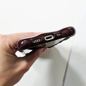 May include: A burgundy phone case with a textured surface. The case has openings for the charging port, speakers, and camera. A black strap is attached to the case. The case is held in a person's hand.