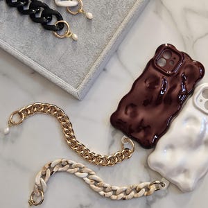 May include: A selection of phone cases and chain accessories. The cases are glossy, in dark brown and white. The chains are gold, black, and cream-coloured, with pearl accents. The items are arranged on a marble surface.