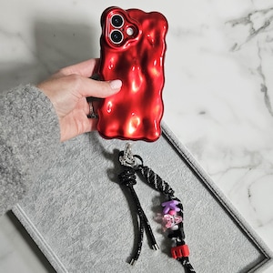 May include: A vibrant red phone case with a distinctive, textured surface. The case is held above a grey tray, with a black phone strap adorned with pink, black, and red beads. The strap is connected to the case with a silver clasp.