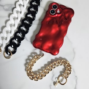 May include: A red, glossy phone case with a wavy design, next to a gold chain bracelet and white and black chain necklaces. The bracelet has a pearl charm. The chains have large links.