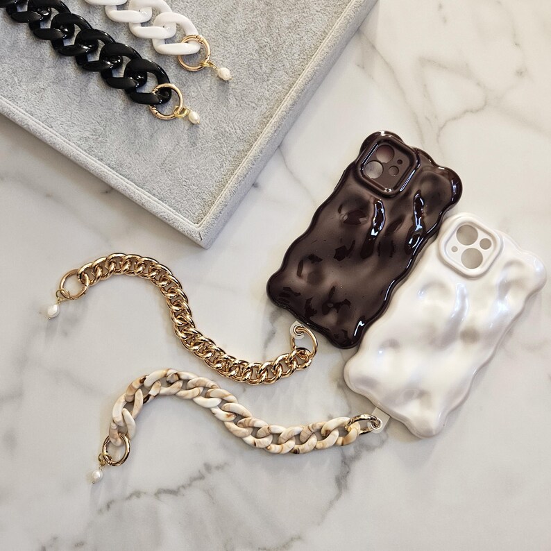 May include: A collection of phone cases and chain straps. The cases are dark brown and white with a textured, wavy design. The chain straps are black, white, gold, and cream-colored, with a pearl accent. The items are displayed on a marble surface.