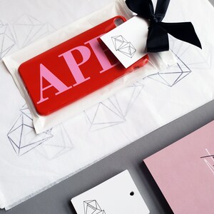 May include: A red phone case with the letters "APH" in pink. The case is packaged in a white tissue paper with a diamond pattern. The case is tied with a black ribbon and a white tag with the words "Diamond Luxe" printed on it.