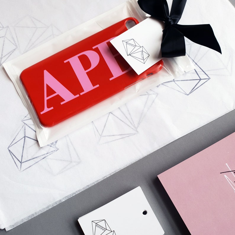 May include: A red phone case with the letters "APH" in pink. The case is packaged in a white tissue paper with a diamond pattern. The case is tied with a black ribbon and a white tag with the words "Diamond Luxe" printed on it.