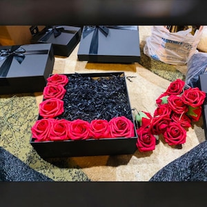 May include: A square black gift box filled with black shredded paper filler and decorated with bright red artificial roses along the edges. Several additional red roses are placed outside the box, and other closed black gift boxes are nearby.