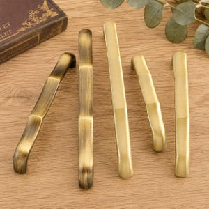 May include: Five metal cabinet pulls arranged in a row. Three pulls feature a brushed antique brass finish with visible vertical grain, while two pulls have a smooth, matte gold finish. Each pull has a long, slender, and slightly curved shape.