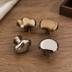 May include: Four cabinet knobs with irregular, rounded shapes. Two knobs feature a brushed brass finish with a matte texture, while the other two knobs have a polished, reflective silver-tone finish. Each knob includes a cylindrical base for mounting.