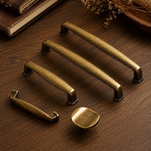 May include: A collection of four antique brass cabinet pulls and one matching square cabinet knob. Each piece features a textured, weathered gold finish with dark highlights, showcasing a curved design with rectangular mounting bases.