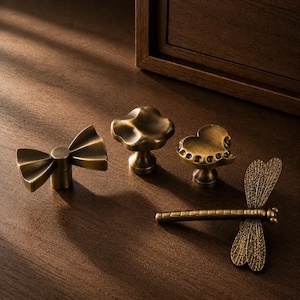 May include: A set of four brass-toned metal cabinet knobs featuring distinct shapes: a bow, a rounded mushroom-like form, a heart with a beaded edge, and a detailed dragonfly with textured wings. Each knob has a central mounting post.