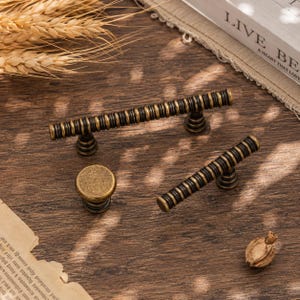 May include: A set of three cabinet hardware pieces in an antique brass finish with dark, recessed grooves. The collection includes one long bar pull, one shorter bar pull, and one round knob, all featuring a consistent ribbed, cylindrical design.